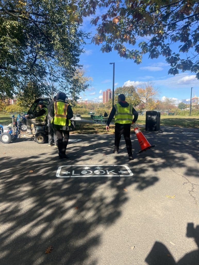 Workers in safety vests stand near a small utility vehicle on a paved park path with trees overhead. A fresh “LOOK” marking with arrows is painted on the pavement, reminding pedestrians to check for cross traffic.