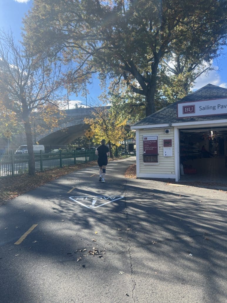 A person jogs on a riverside path beside the BU Sailing Pavilion building. The word “SLOW!” is freshly painted on the asphalt ahead, surrounded by autumn trees and sunlight filtering through the branches.