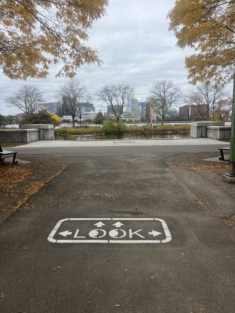 A paved path leading toward a river with the Cambridge skyline visible in the distance. The word “LOOK” is painted on the pavement with arrows pointing left, right, and straight ahead. Trees with yellow autumn leaves frame the view.