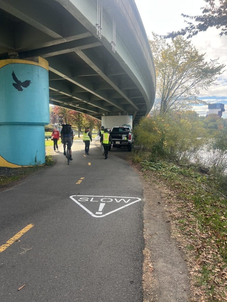People wearing safety vests walk and cycle under a bridge on a paved river path. A “SLOW!” marking is painted on the pavement, and a pickup truck is parked ahead near a bright blue bridge column with a bird silhouette.