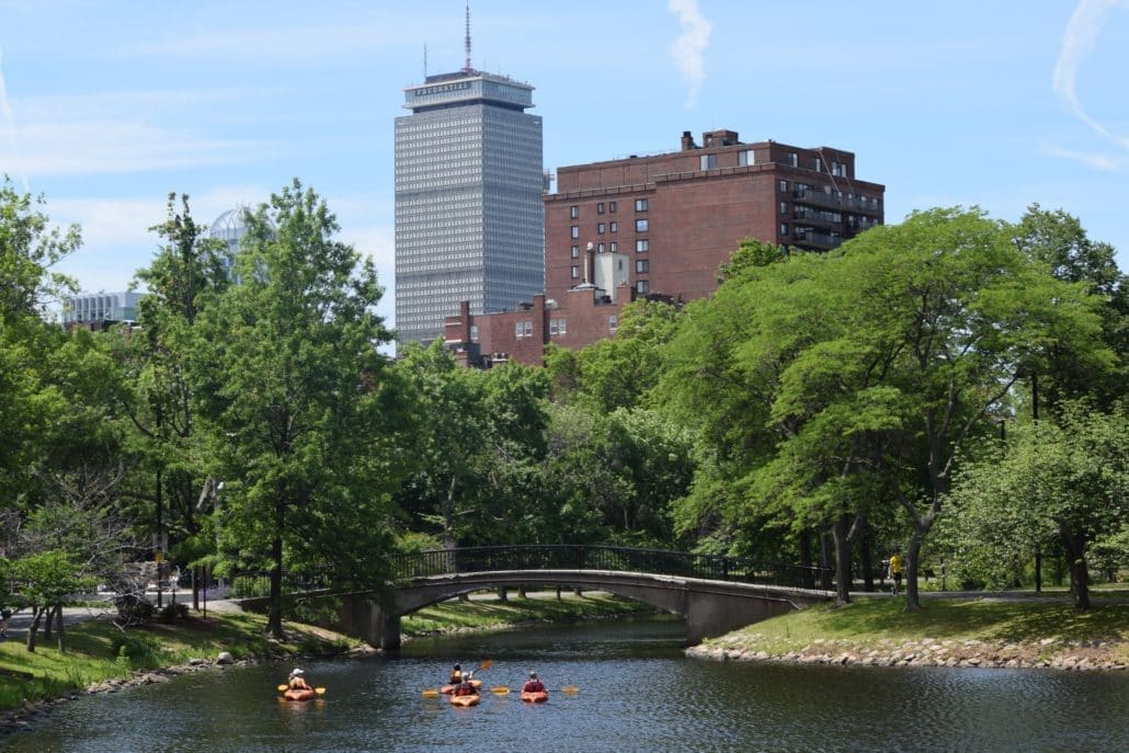 The Charles River Esplanade — kayakers paddle past an arched footbridge with the Boston skyline beyond.