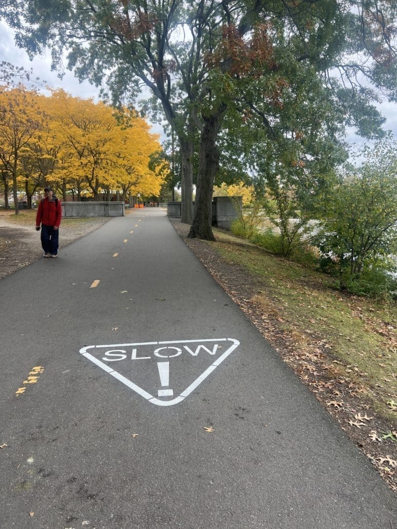 A paved riverside path lined with trees in bright fall colors. The word “SLOW” with an exclamation mark inside a white triangle is painted on the pavement. A person in a red jacket walks along the path.