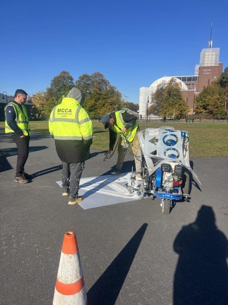 Workers in bright yellow safety vests use a stencil and painting machine to mark a symbol on a paved path near the Museum of Science. An orange traffic cone is placed in the foreground, and autumn trees and a clear blue sky are visible in the background.