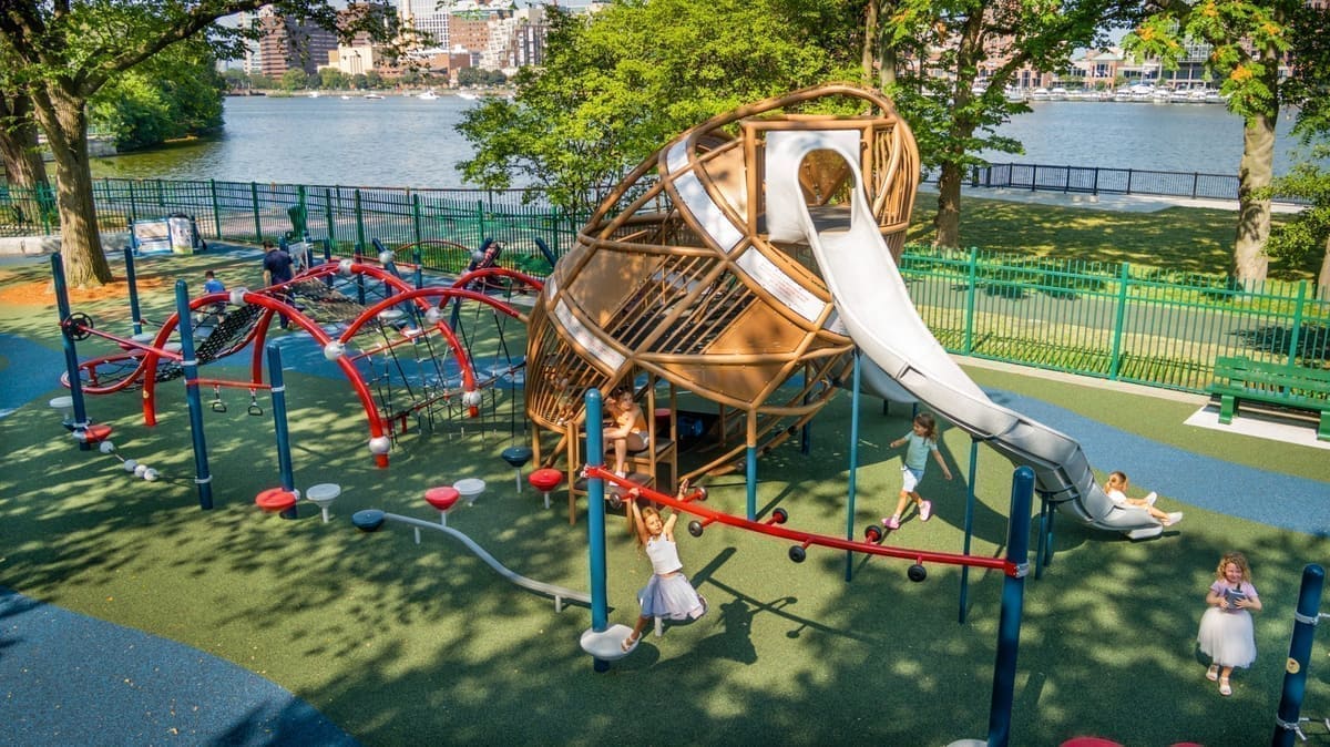 Children play on the riverside Charlesbank playground with the Charles River and Boston skyline behind.